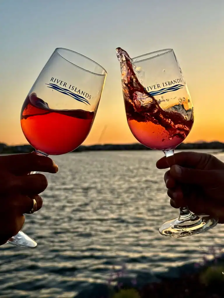 Two wine glasses clink in a sunset toast over a lake with River Islands branding on the glasses